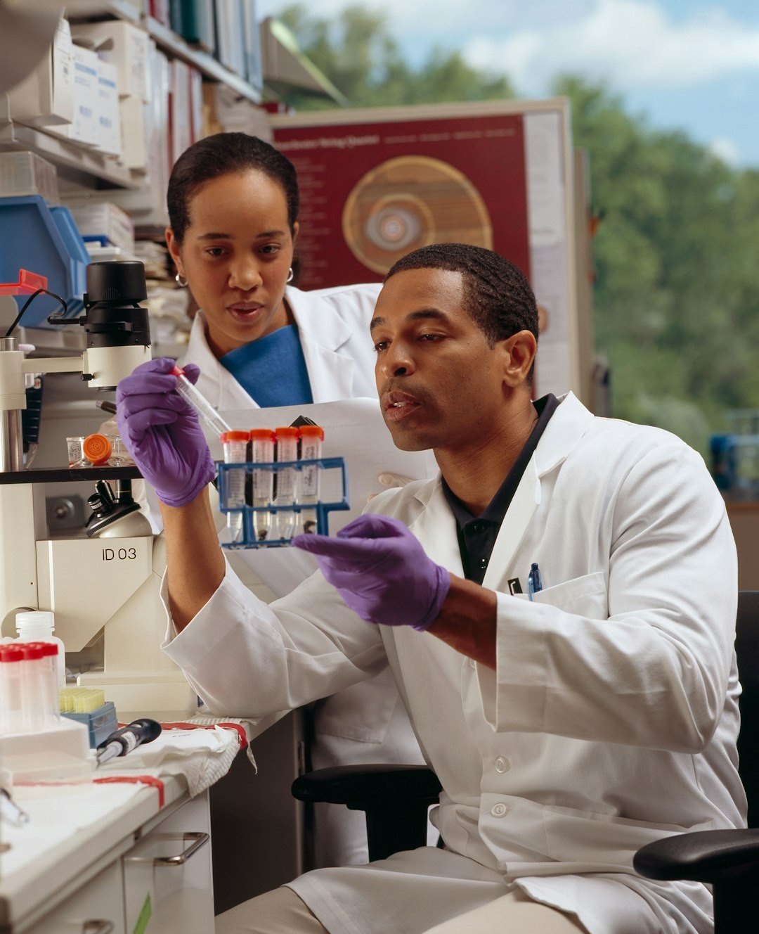 Researcher Checks Test Tubes. An African American male researcher checks test tubes as an AfricanAmerican female cancer researcher looks on.